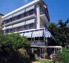 albergo visto di fronte, viale di ingresso, entrata, soggiorno, terrazzi delle camere con le tende a capottina, giardino, sullo sfondo altri alberghi di Riccione